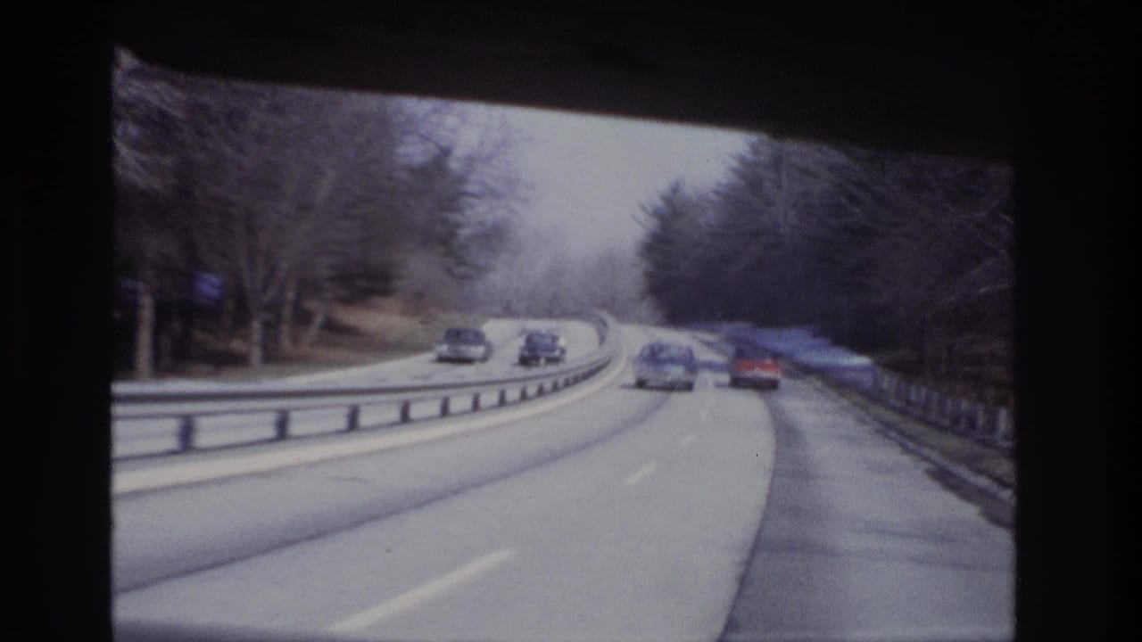 1960s home-movie driving view with multiple cars on a curved roadway, bare trees, guardrails, and soft 8mm color.