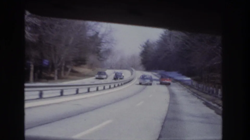 1960s home-movie driving view with multiple cars on a curved roadway, bare trees, guardrails, and soft 8mm color.