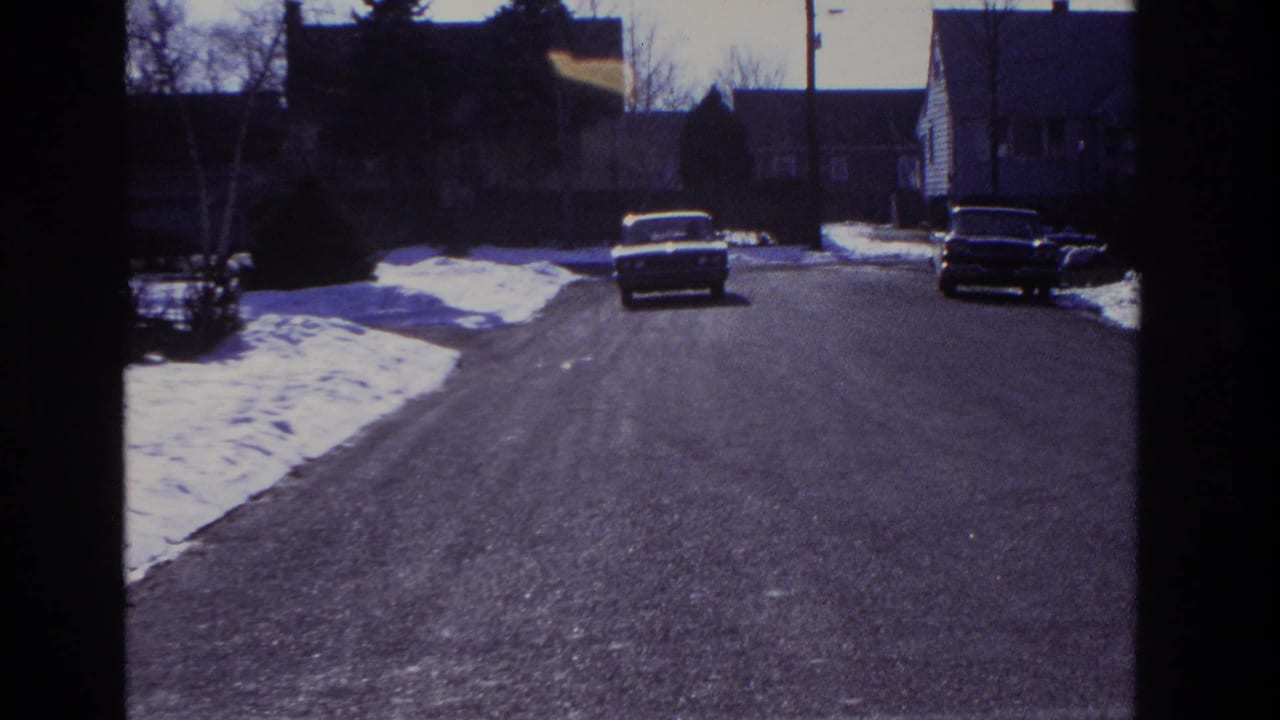 The driving view shifts to a quiet residential street with snow piles, parked classic cars, and houses visible ahead.