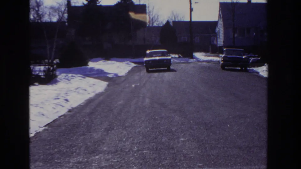 The driving view shifts to a quiet residential street with snow piles, parked classic cars, and houses visible ahead.