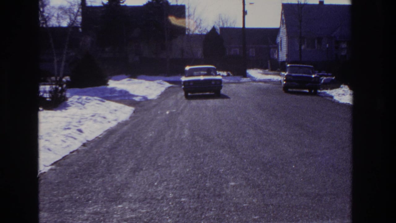 A light-colored sedan approaches along a snow-lined residential street in the supplied 1966 Hoboken home-movie footage.