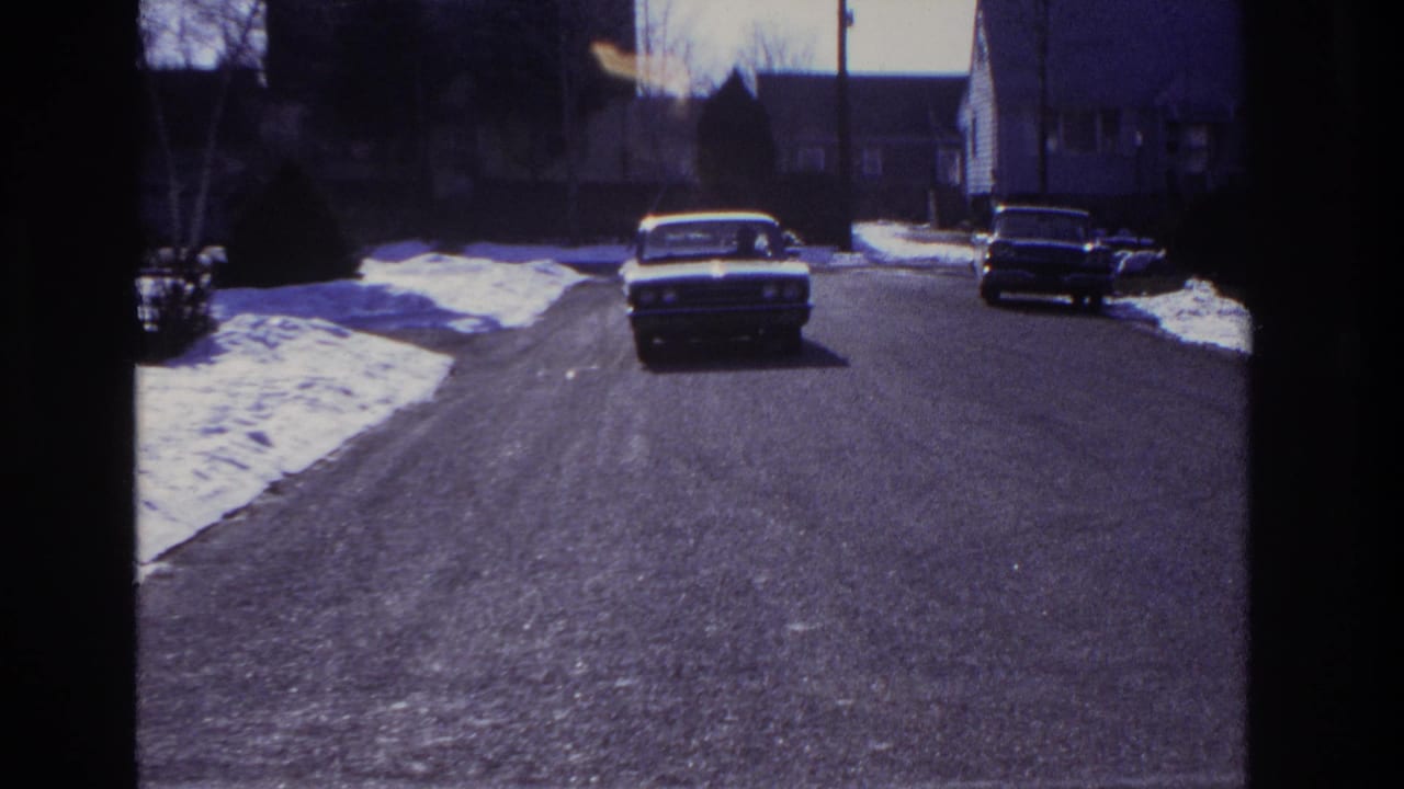 The sedan advances toward camera on a quiet Hoboken street, with snow patches and parked cars framing the roadway.