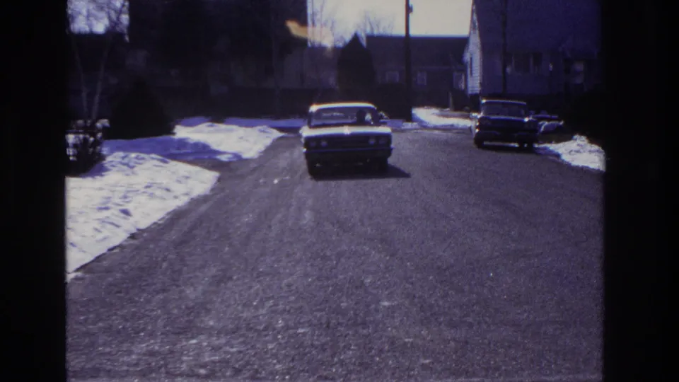 The sedan advances toward camera on a quiet Hoboken street, with snow patches and parked cars framing the roadway.