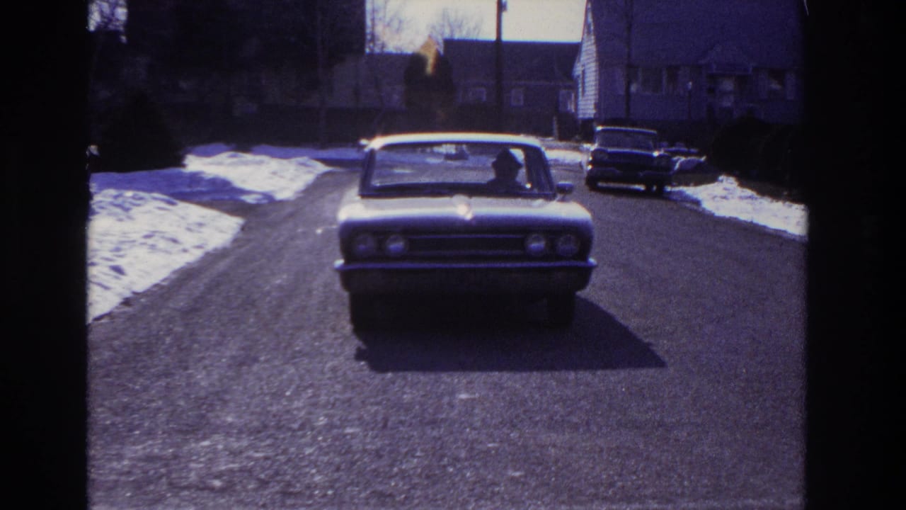 The sedan reaches the foreground on the Hoboken street, with a driver silhouette visible through the windshield.