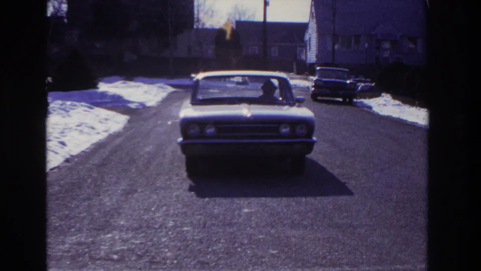 The sedan reaches the foreground on the Hoboken street, with a driver silhouette visible through the windshield.