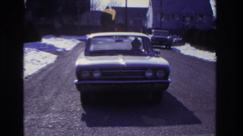 1966 Hoboken home-movie frame of a light-colored sedan approaching on a quiet snow-lined residential street.