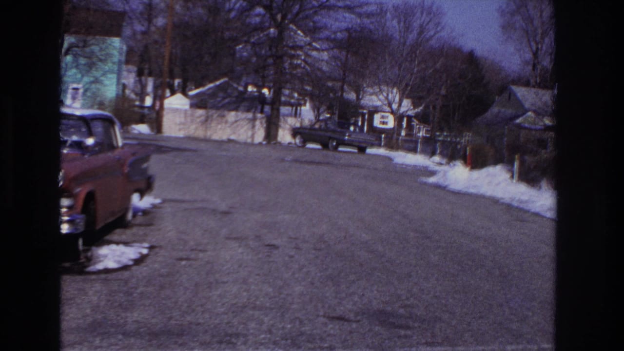 Residential street with parked cars, snow patches, houses and bare trees.