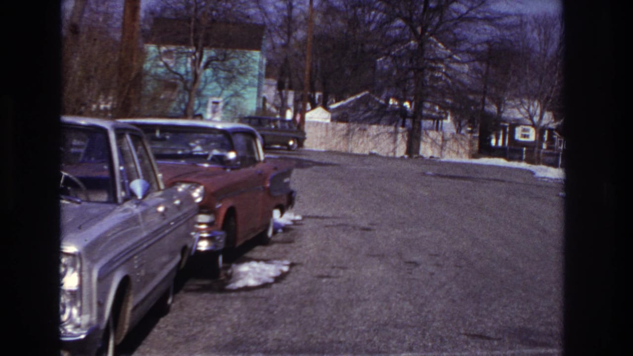 1966 Hoboken home-movie frame with parked mid-century sedans, snow patches, bare trees, houses, and a dark car up the street.