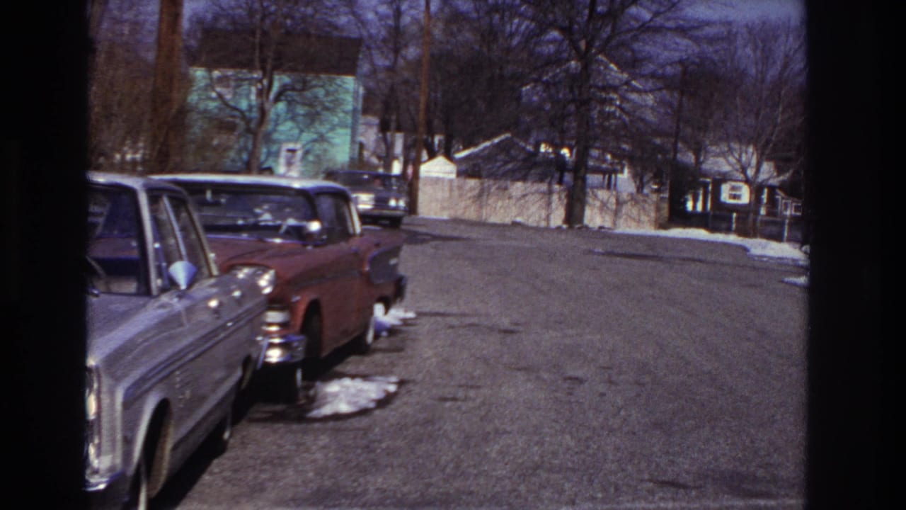A dark sedan moves near the far curb on a Hoboken residential street, with parked 1960s cars, bare trees, snow patches, and houses visible.