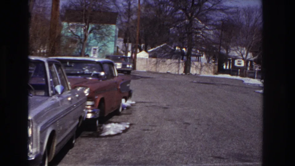 A dark sedan moves near the far curb on a Hoboken residential street, with parked 1960s cars, bare trees, snow patches, and houses visible.