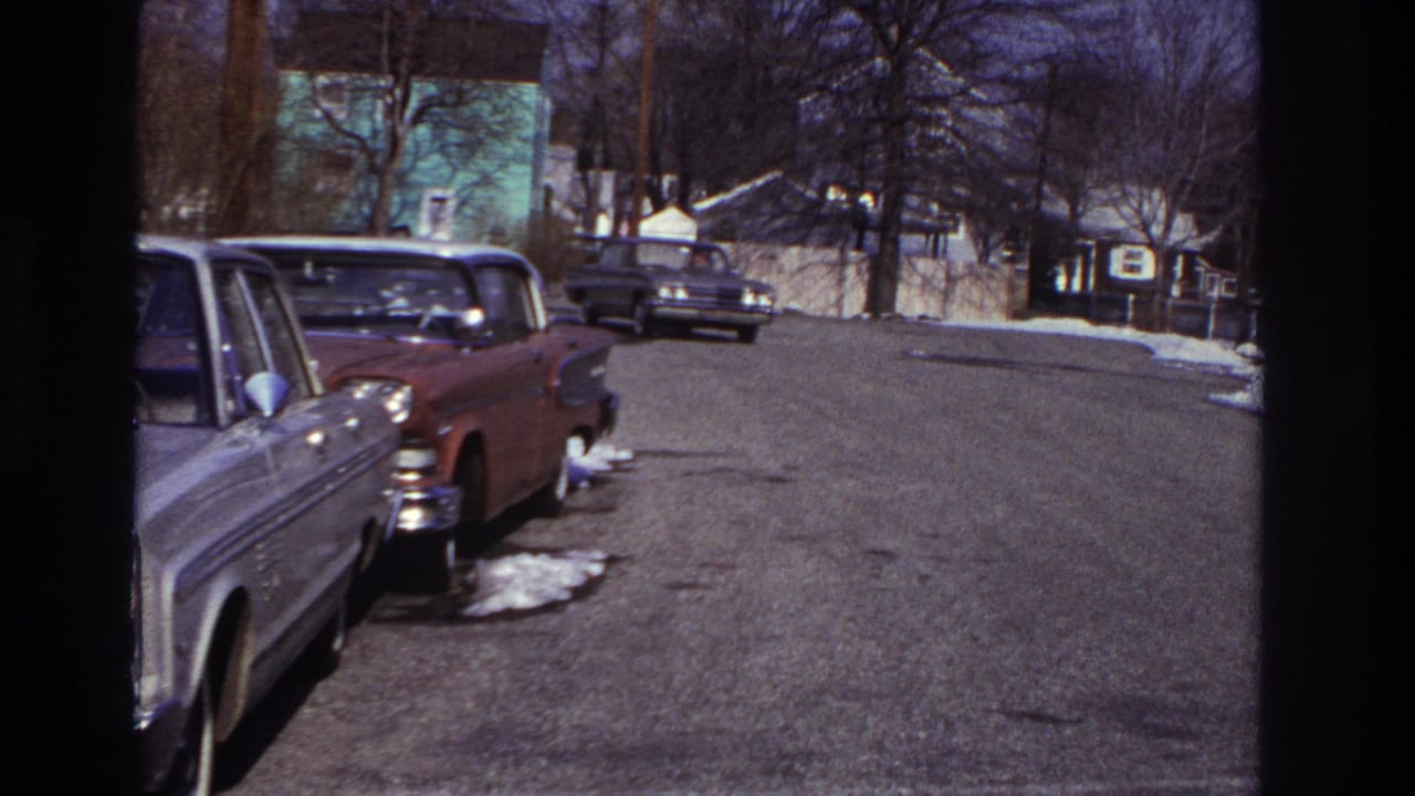 Mid-turn view of a dark sedan on a Hoboken residential block, with parked cars, winter snow patches, leafless trees, and small distant people.