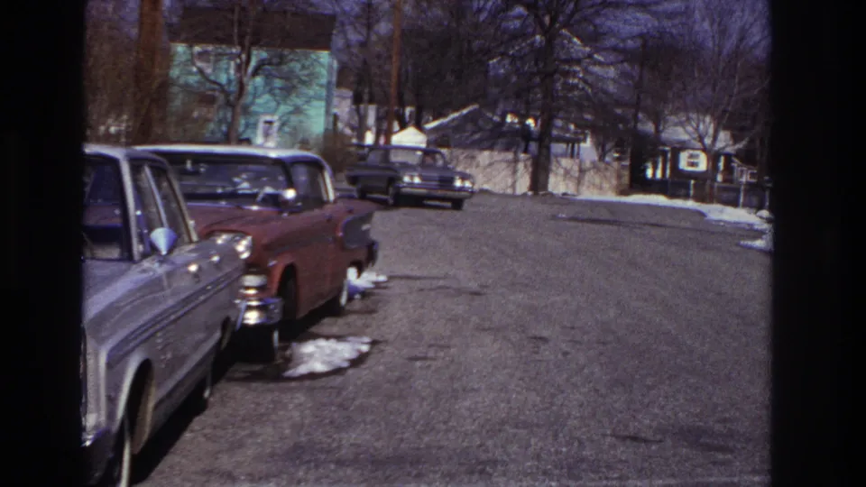 Mid-turn view of a dark sedan on a Hoboken residential block, with parked cars, winter snow patches, leafless trees, and small distant people.
