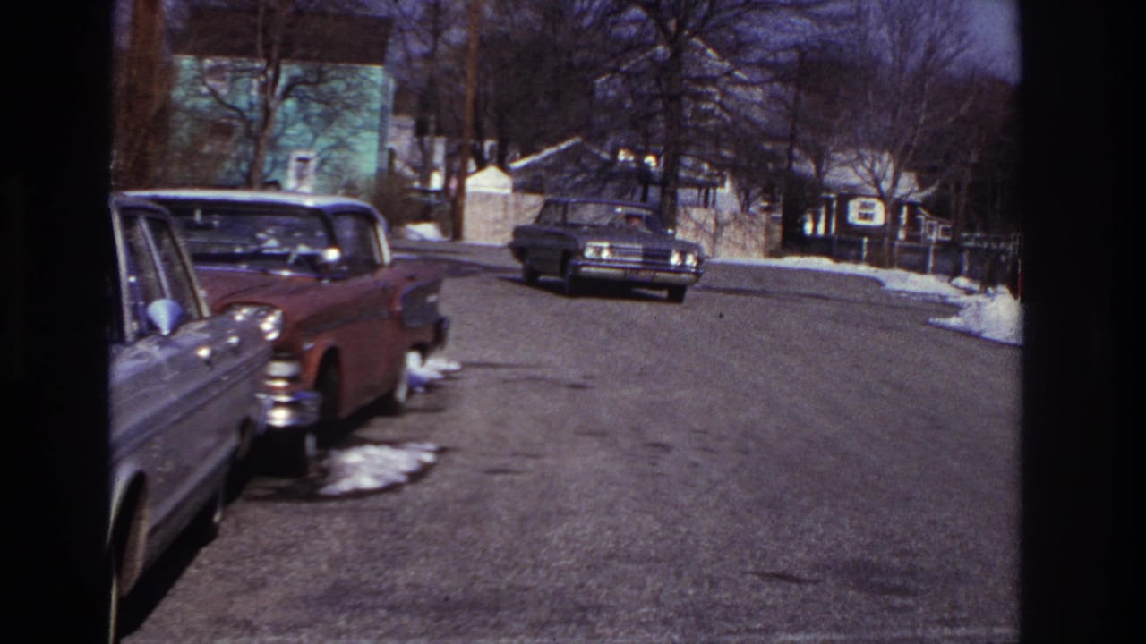 The dark sedan completes its slow turn on a 1966 Hoboken residential street, passing parked mid-century cars, bare trees, fences, and winter snow remnants.