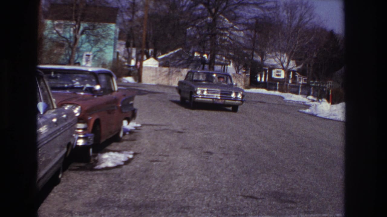 1966 Hoboken home-movie frame showing a quiet winter residential street with parked cars, snow patches, houses, and a distant approaching sedan.