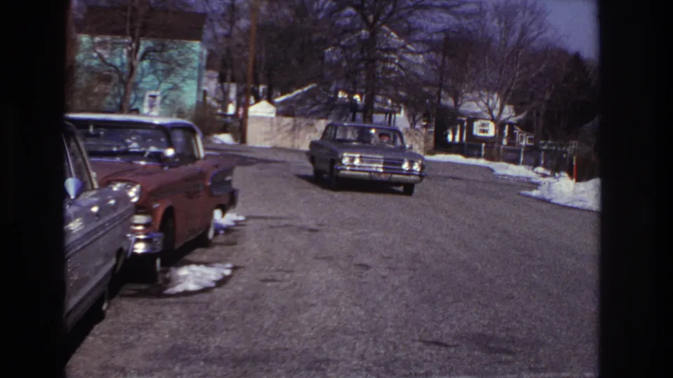 1966 Hoboken home-movie frame showing a quiet winter residential street with parked cars, snow patches, houses, and a distant approaching sedan.