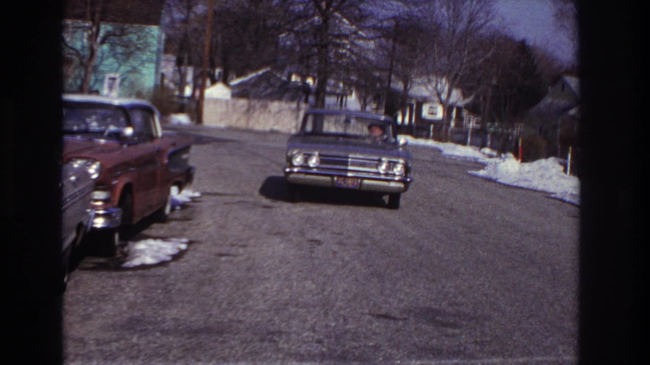Classic sedan driving toward the camera on a residential street with snow along the curb.