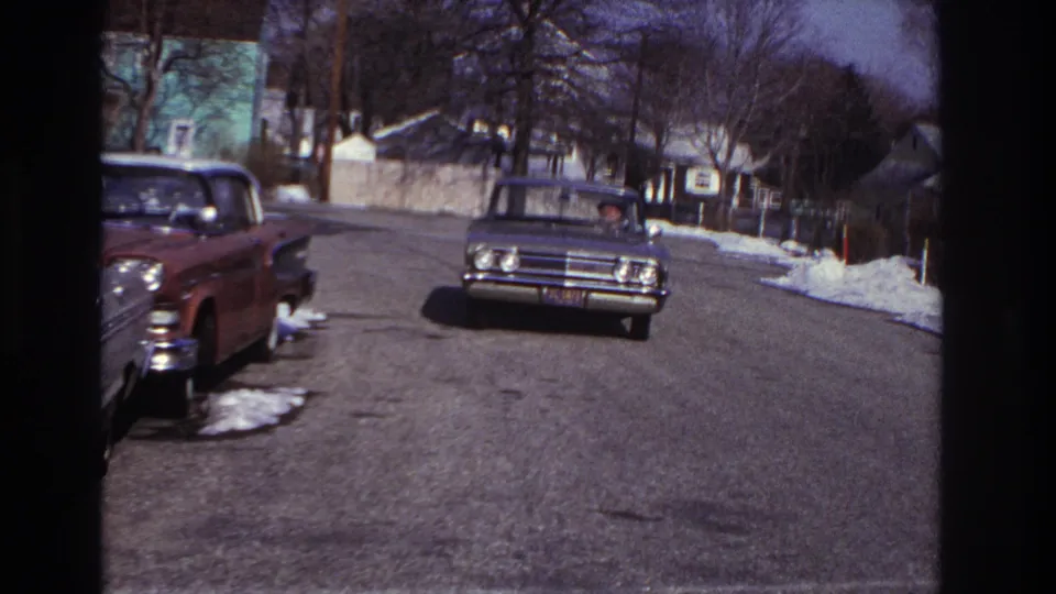 1966 Hoboken street view with a gray classic sedan nearing the camera, parked cars at left, winter trees, houses, and remaining snow on the curb.