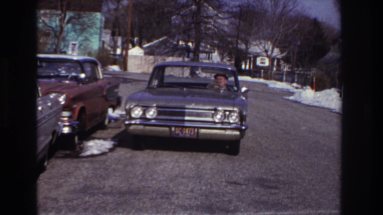 1966 Hoboken home-movie frame showing a classic sedan near the camera, driver visible, parked cars, snow patches, and houses on a winter street.