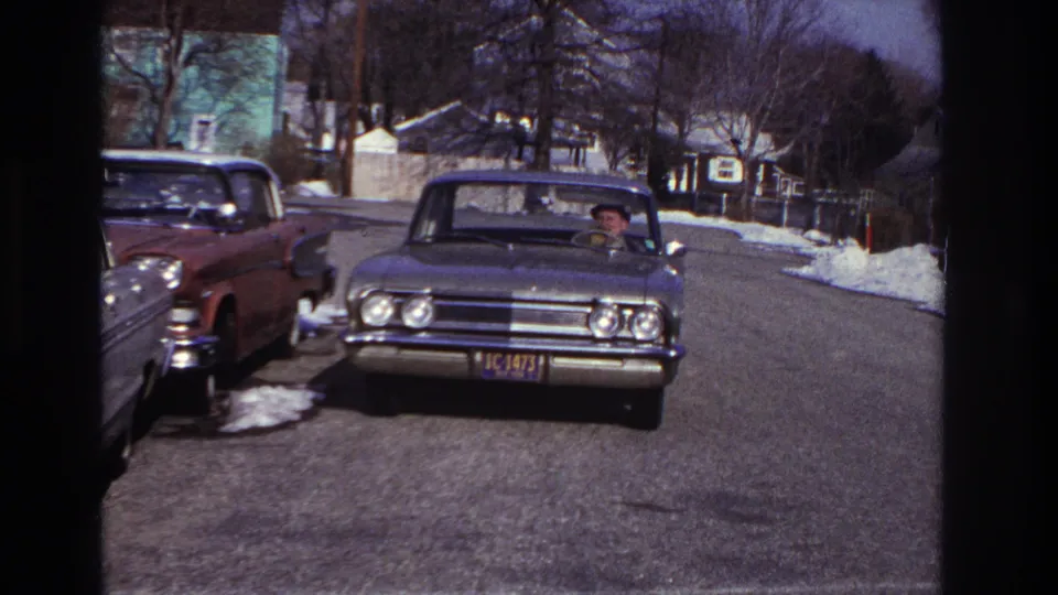 1966 Hoboken home-movie frame showing a classic sedan near the camera, driver visible, parked cars, snow patches, and houses on a winter street.