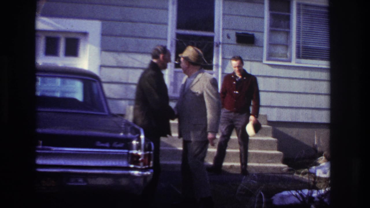The 1966 Hoboken home movie shows two adult men in close greeting near a dark sedan, with a third man holding a hat near the house steps.