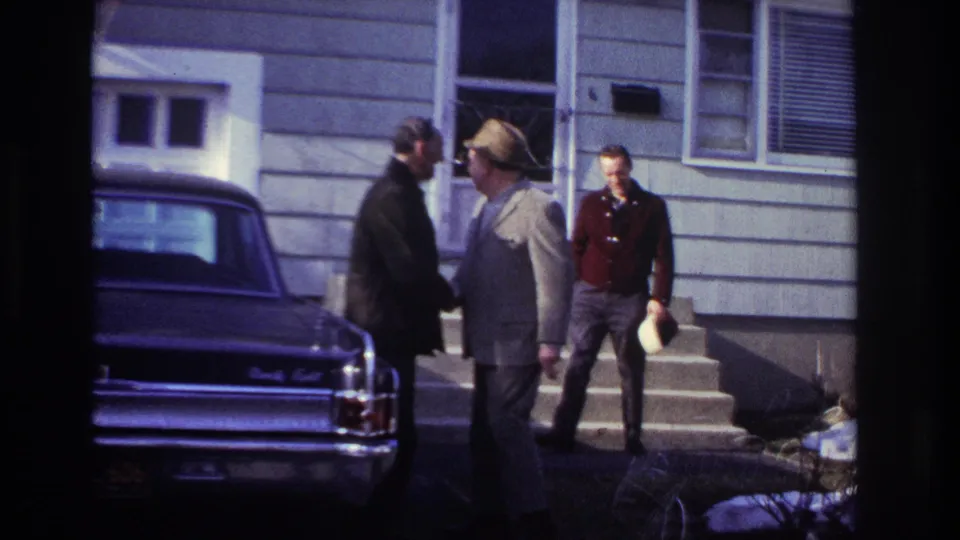 The 1966 Hoboken home movie shows two adult men in close greeting near a dark sedan, with a third man holding a hat near the house steps.