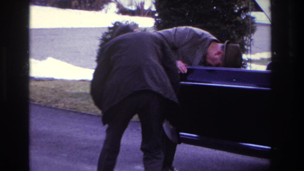 A man in a coat and hat bends over the rear of a dark sedan on a driveway, with snow patches visible in the yard.