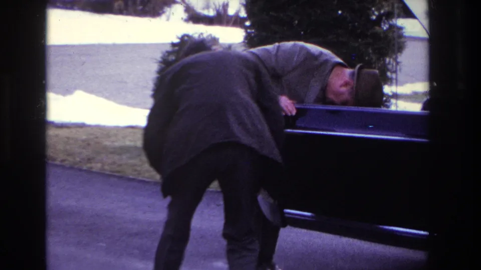 A man in a coat and hat bends over the rear of a dark sedan on a driveway, with snow patches visible in the yard.