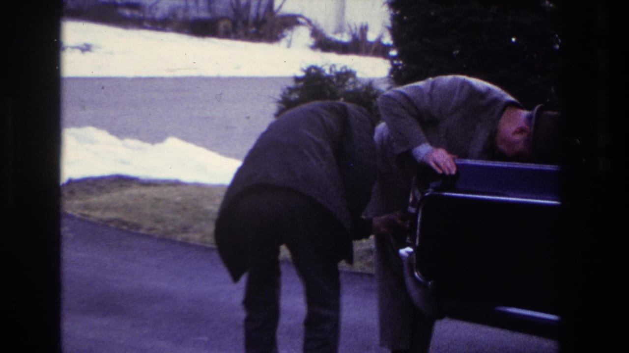 In 1966 Hoboken home-movie footage, two men in coats and hats bend over an open car on a driveway bordered by snow.