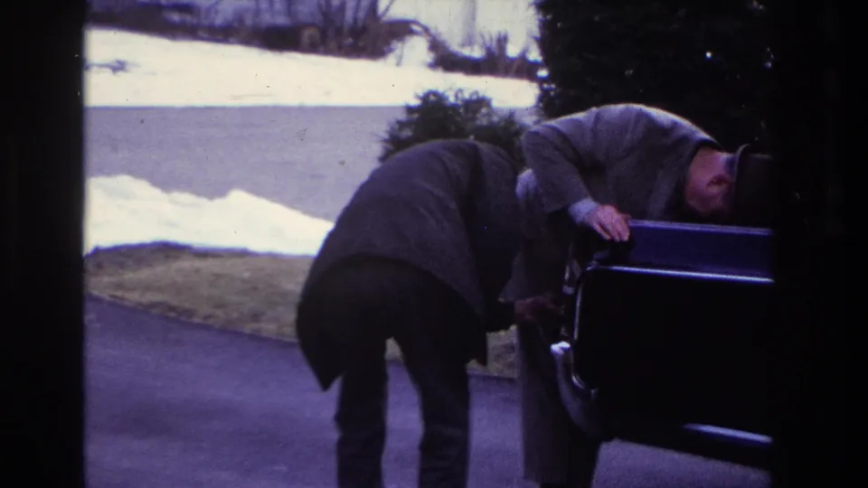 In 1966 Hoboken home-movie footage, two men in coats and hats bend over an open car on a driveway bordered by snow.