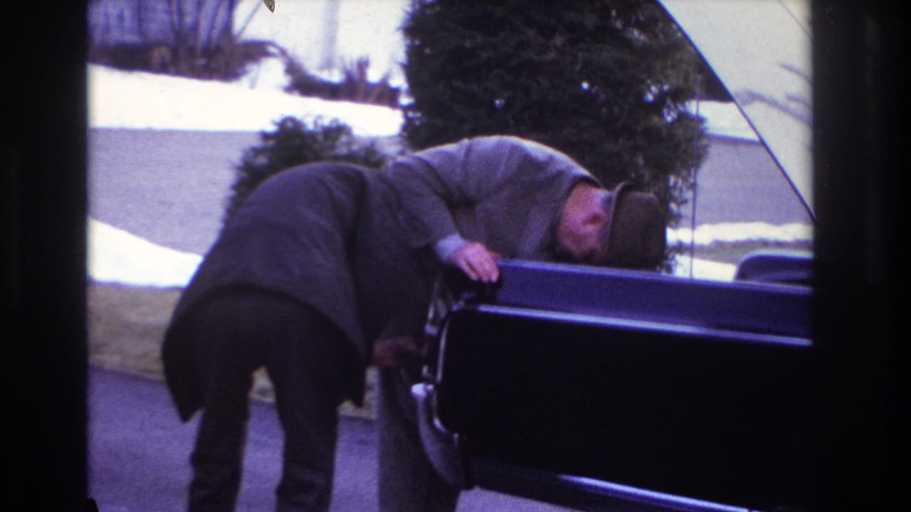 The 8mm frame shows two men closely inspecting or working at an open car on a residential driveway in Hoboken.