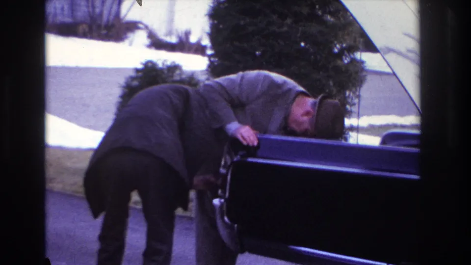 The 8mm frame shows two men closely inspecting or working at an open car on a residential driveway in Hoboken.