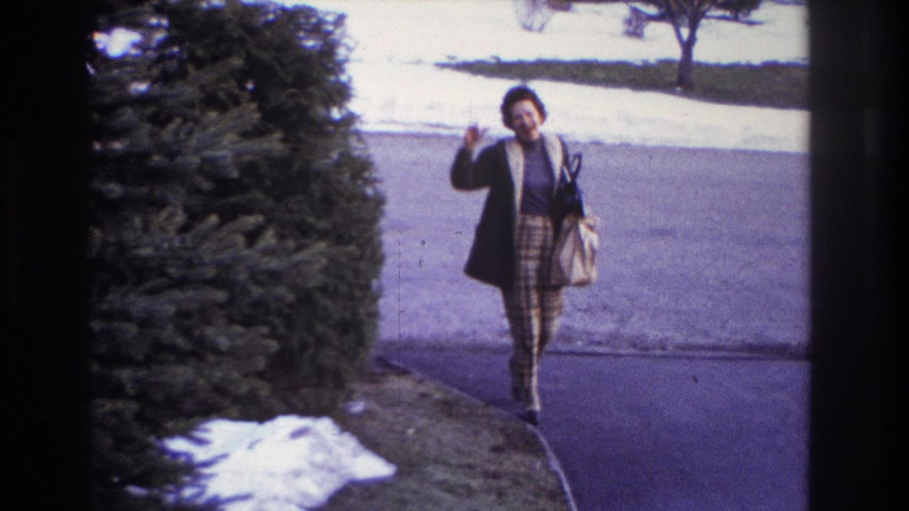 A woman in a coat and patterned skirt walks toward the home-movie camera in Hoboken, carrying a handbag near the driveway.