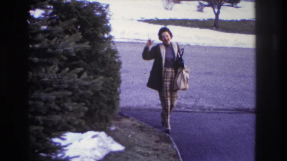 A woman in a coat and patterned skirt walks toward the home-movie camera in Hoboken, carrying a handbag near the driveway.