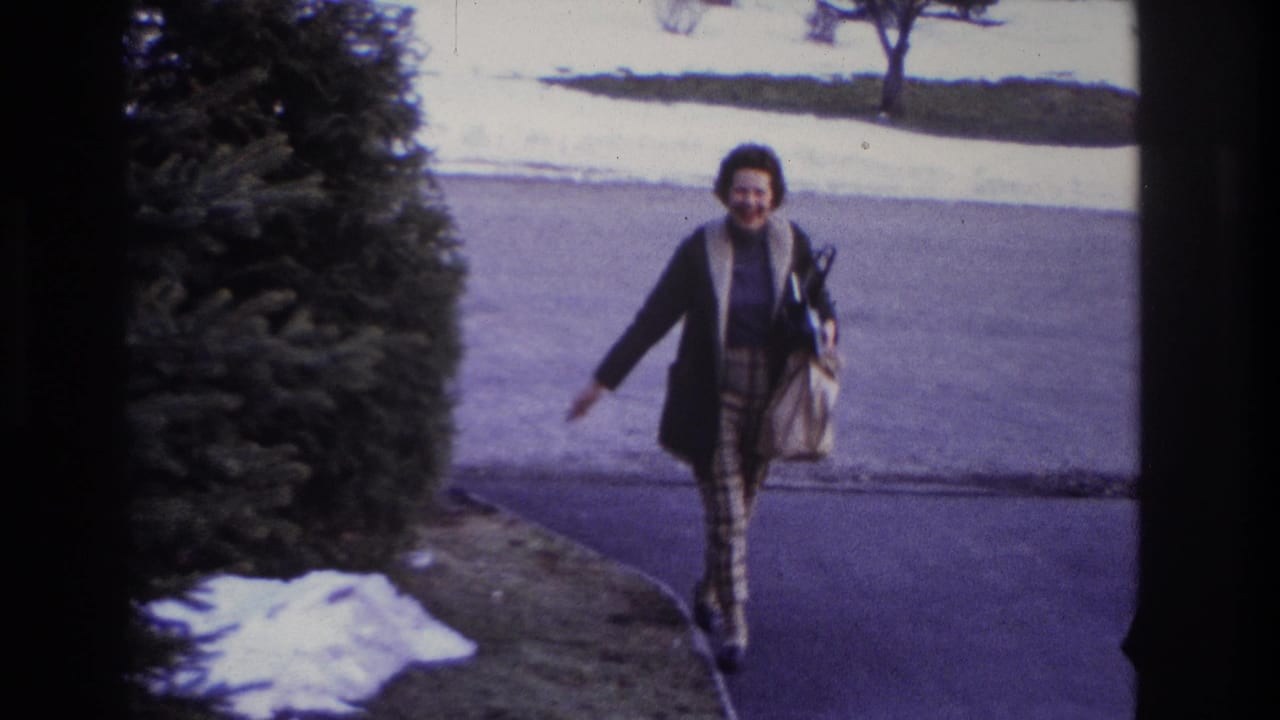 In 1966 Hoboken home-movie footage, a smiling woman walks down a residential driveway with a handbag, snow patches, and an evergreen shrub nearby.