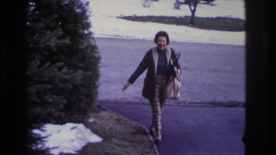 In 1966 Hoboken home-movie footage, a smiling woman walks down a residential driveway with a handbag, snow patches, and an evergreen shrub nearby.