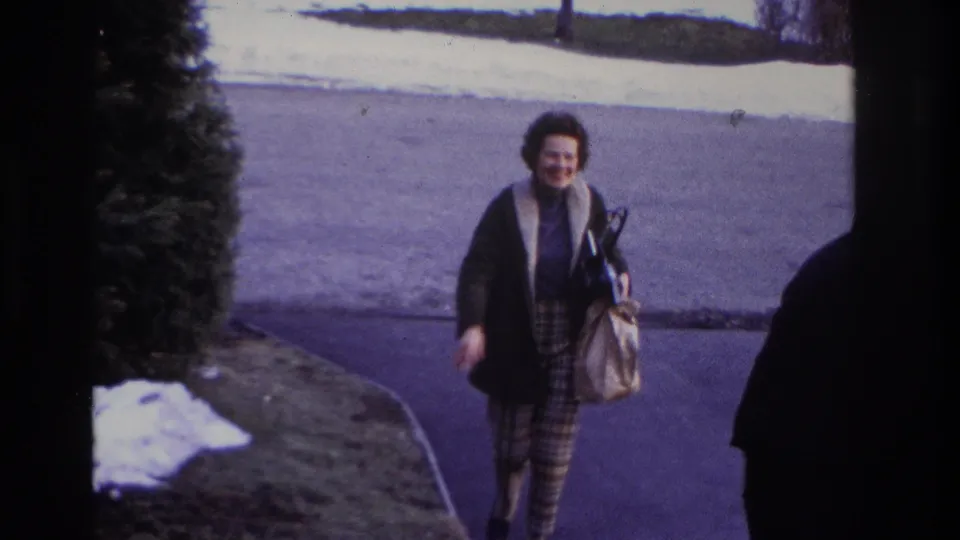 The woman moves closer in the Hoboken driveway scene, smiling with a handbag while snow and a dark vehicle edge appear near the frame.