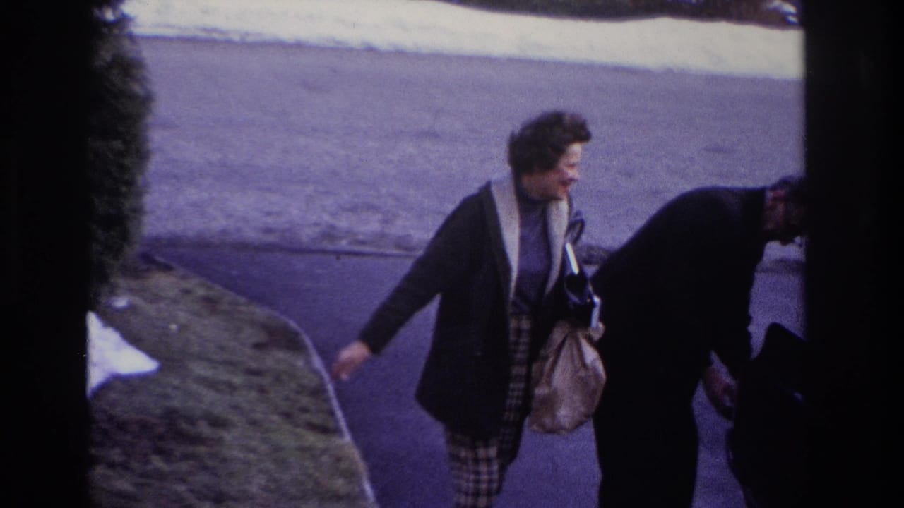 The woman passes close to another adult near the driveway, still carrying her handbag as the home-movie camera frames snow and pavement.