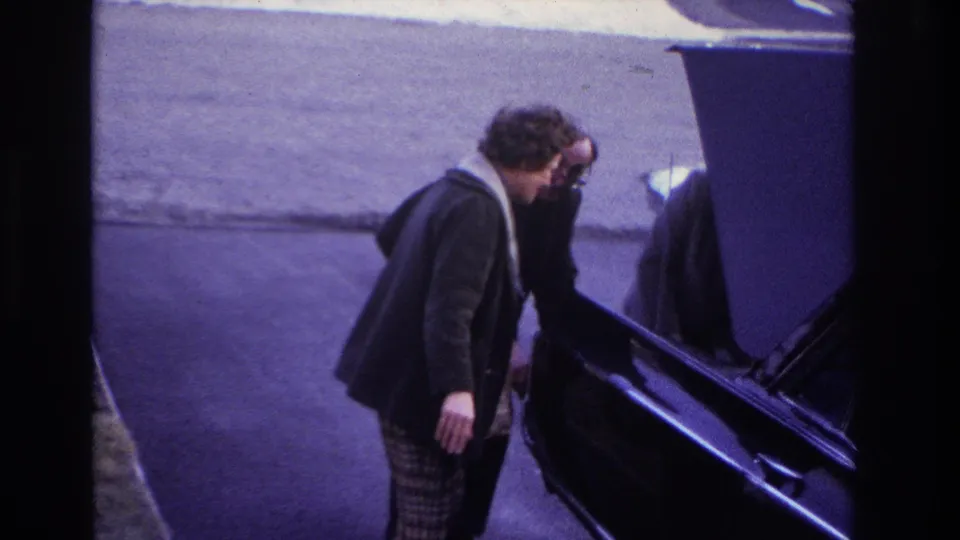 At the end of the 1966 Hoboken clip, the woman stands beside another adult near a dark car, with driveway pavement and snow in view.