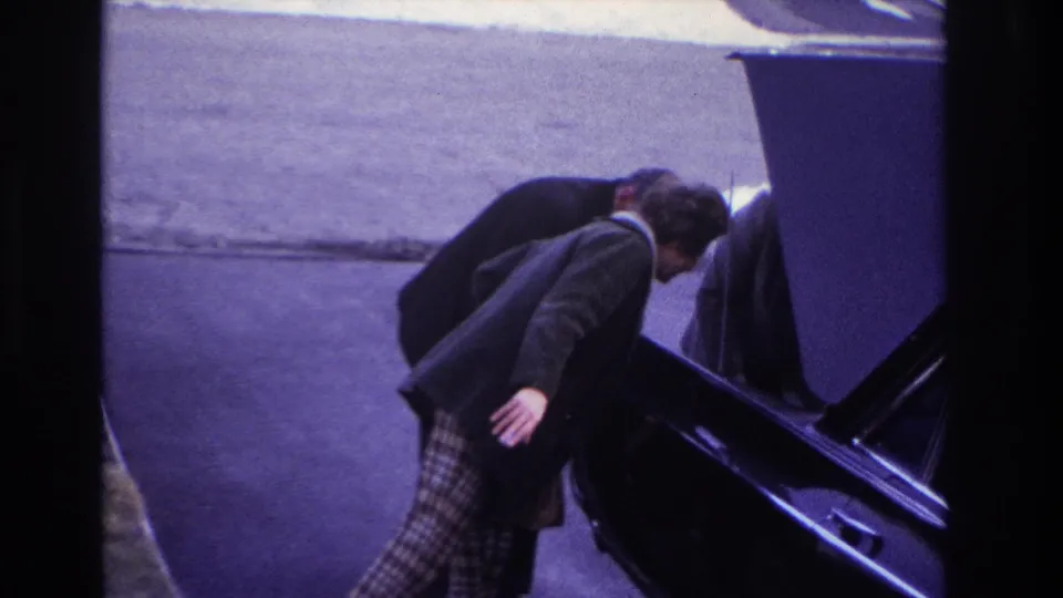 In 1966 Hoboken home-movie footage, a woman in plaid pants stands beside a black car as a man works near the raised hood.