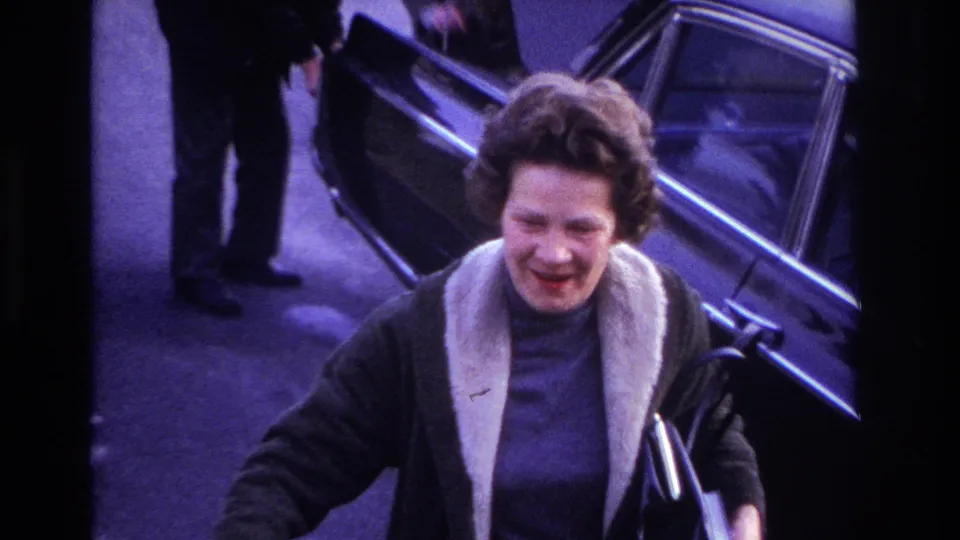 The woman smiles close to the camera near the open black car, with a man and the raised hood visible in the background.