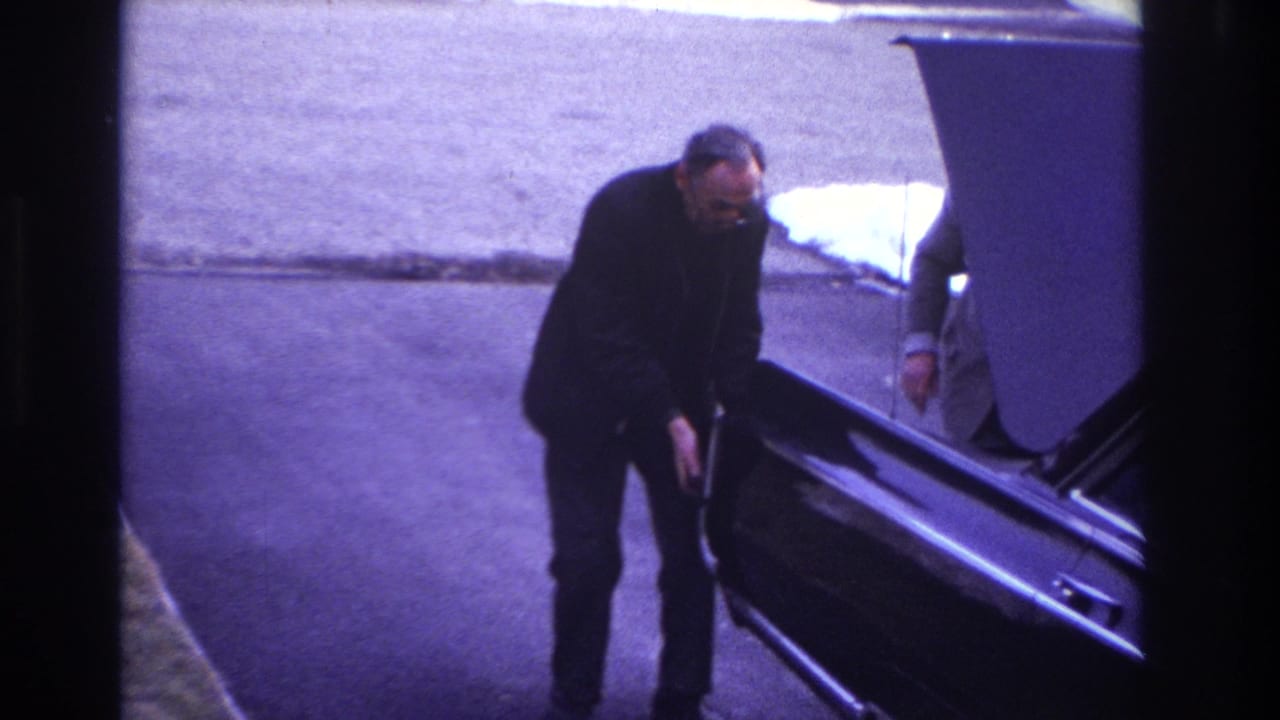 1966 Hoboken home-movie frame showing a suited man pushing a dark sedan with its hood open while another adult is partly visible nearby.