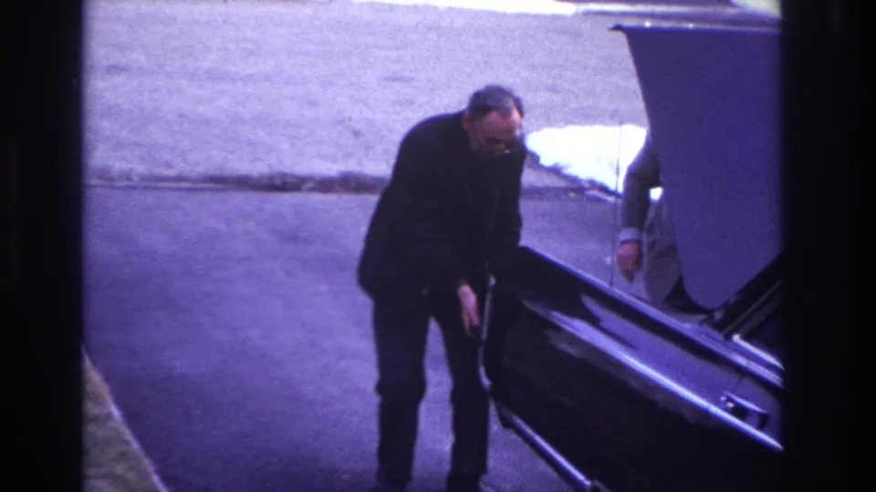 1966 Hoboken home-movie frame showing a suited man pushing a dark sedan with its hood open while another adult is partly visible nearby.