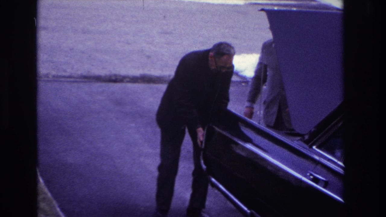 Man bends at the side of a dark car as another suited adult stands near the open hood.