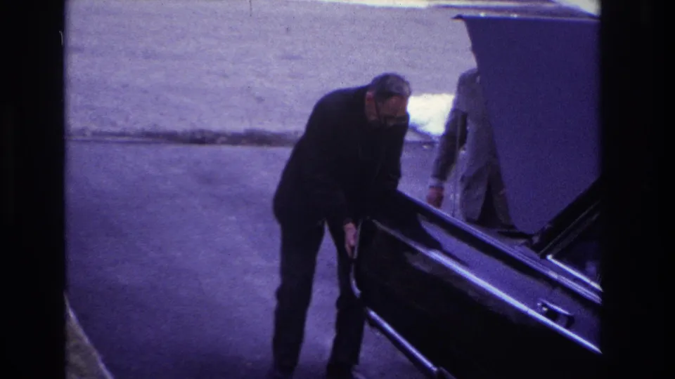 1966 Hoboken frame with a man pushing or steadying a dark car while a second adult in a light suit stands by the raised hood.