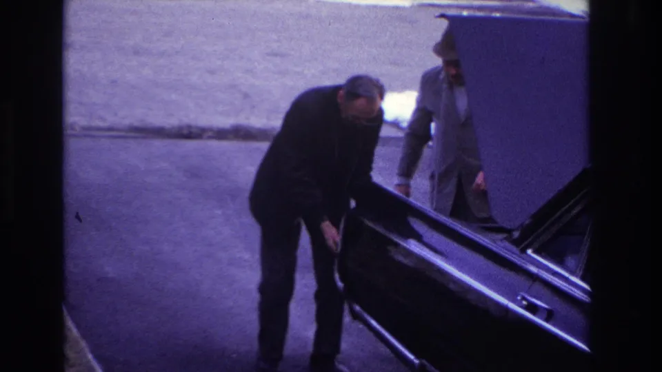 1966 Hoboken home-movie frame showing roadside car trouble as a suited man pushes a sedan and a woman in a dark coat walks past.
