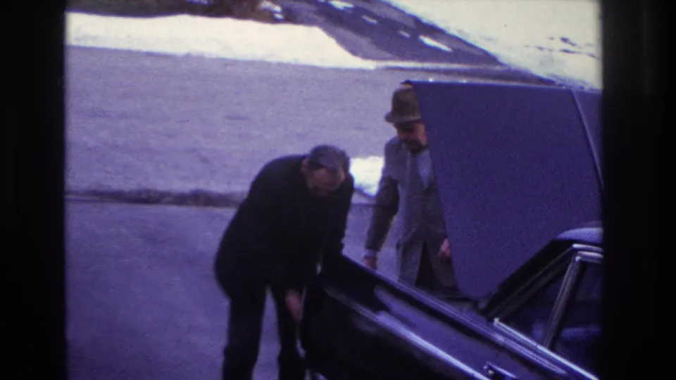 1966 Hoboken frame showing a dark sedan with its hood raised while a man leans at the side and another man in a hat stands nearby.