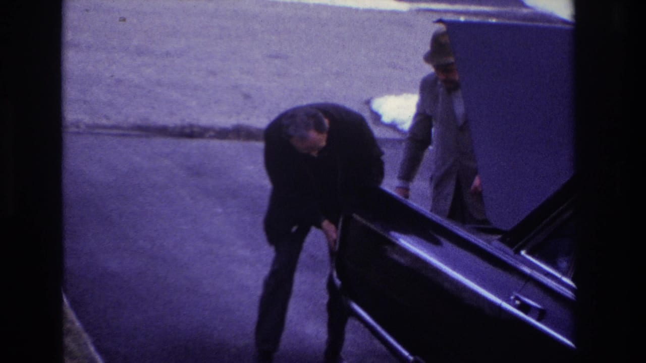 1966 Hoboken home-movie frame showing a man in a dark suit bending near the open trunk of a black car while another man stands nearby.