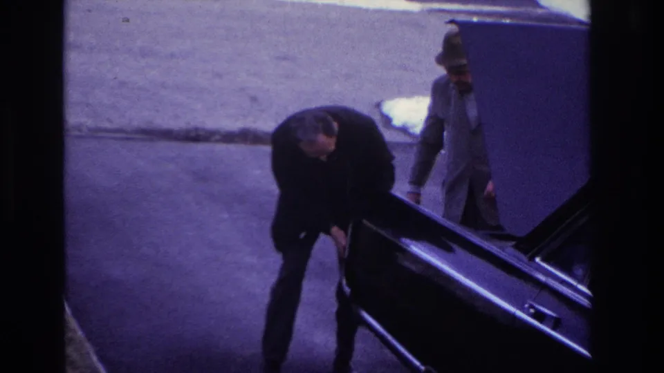 1966 Hoboken home-movie frame showing a man in a dark suit bending near the open trunk of a black car while another man stands nearby.