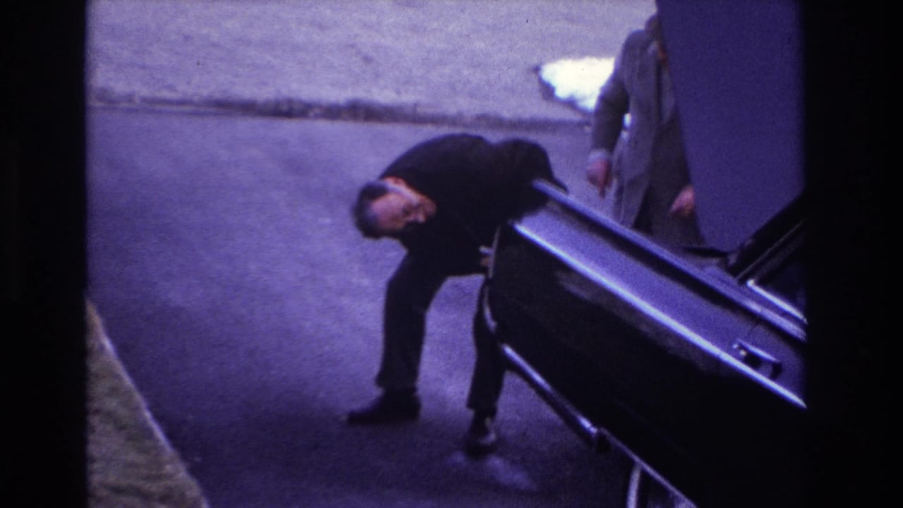 A suited man bends beside the open trunk of a black car.