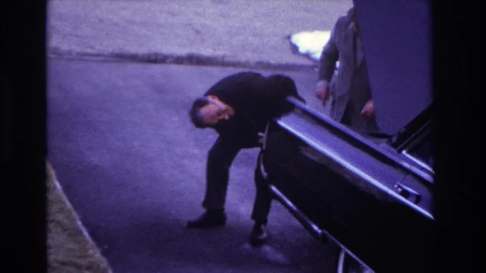 1966 Hoboken frame with a suited man leaning down at the rear of a black car as another adult is partly visible near the open trunk.